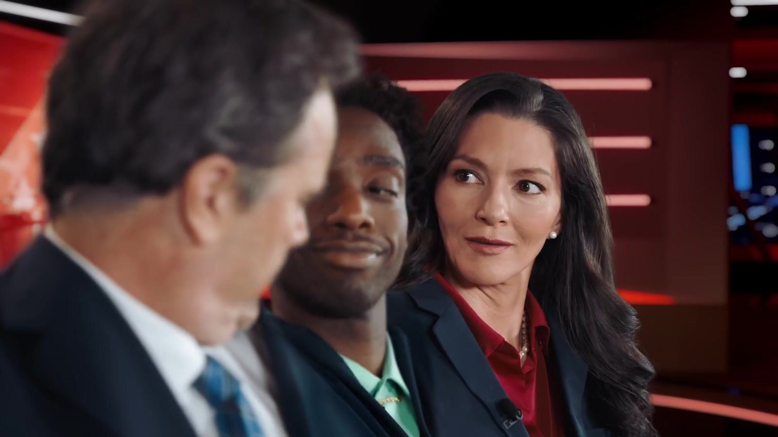 Close shot at the anchor desk with the two anchors and a third performer, red set lighting behind them