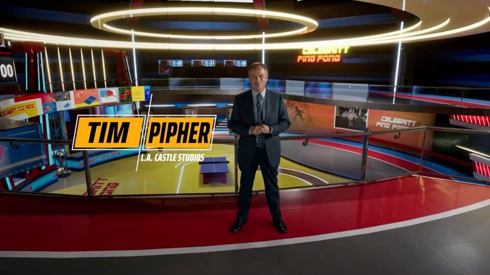 Game show set — host in a suit on a circular 'Celebrity Ping Pong' arena ringed by video screens, a virtual production set at L.A. & ABQ Castle Studios