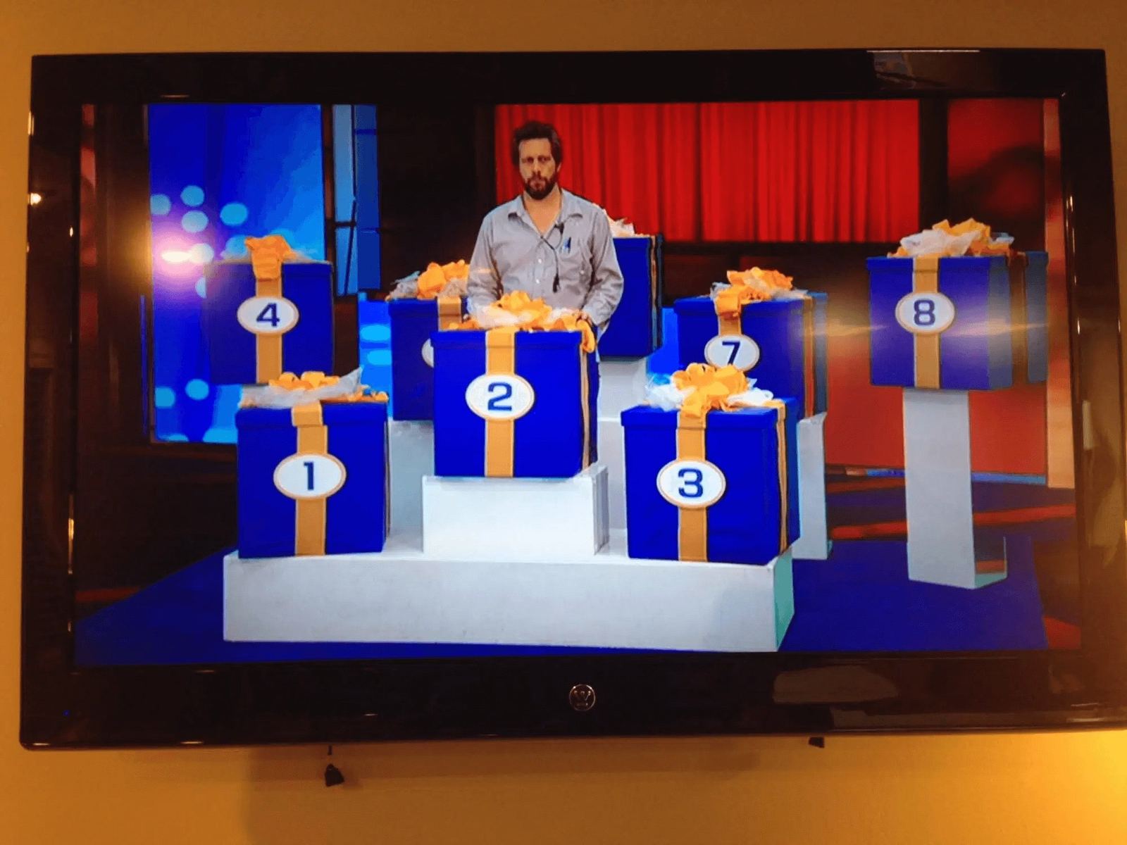 Game show set — a contestant behind numbered blue gift boxes on a prize-reveal podium shown on a monitor at L.A. & ABQ Castle Studios