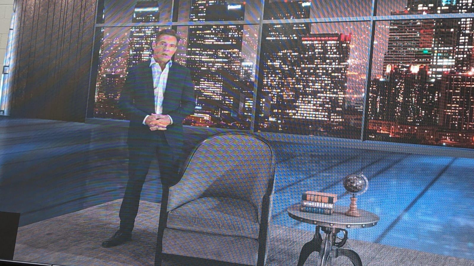 Host wrap: a presenter stands by an accent chair on a virtual set with a nighttime city-skyline window, a virtual production set at L.A. & ABQ Castle Studios