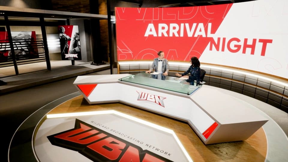 Sports news set — two WBN broadcasters at an angular desk on a circular floor with a red 'Arrival Night' video wall, a virtual production set at L.A. & ABQ Castle Studios