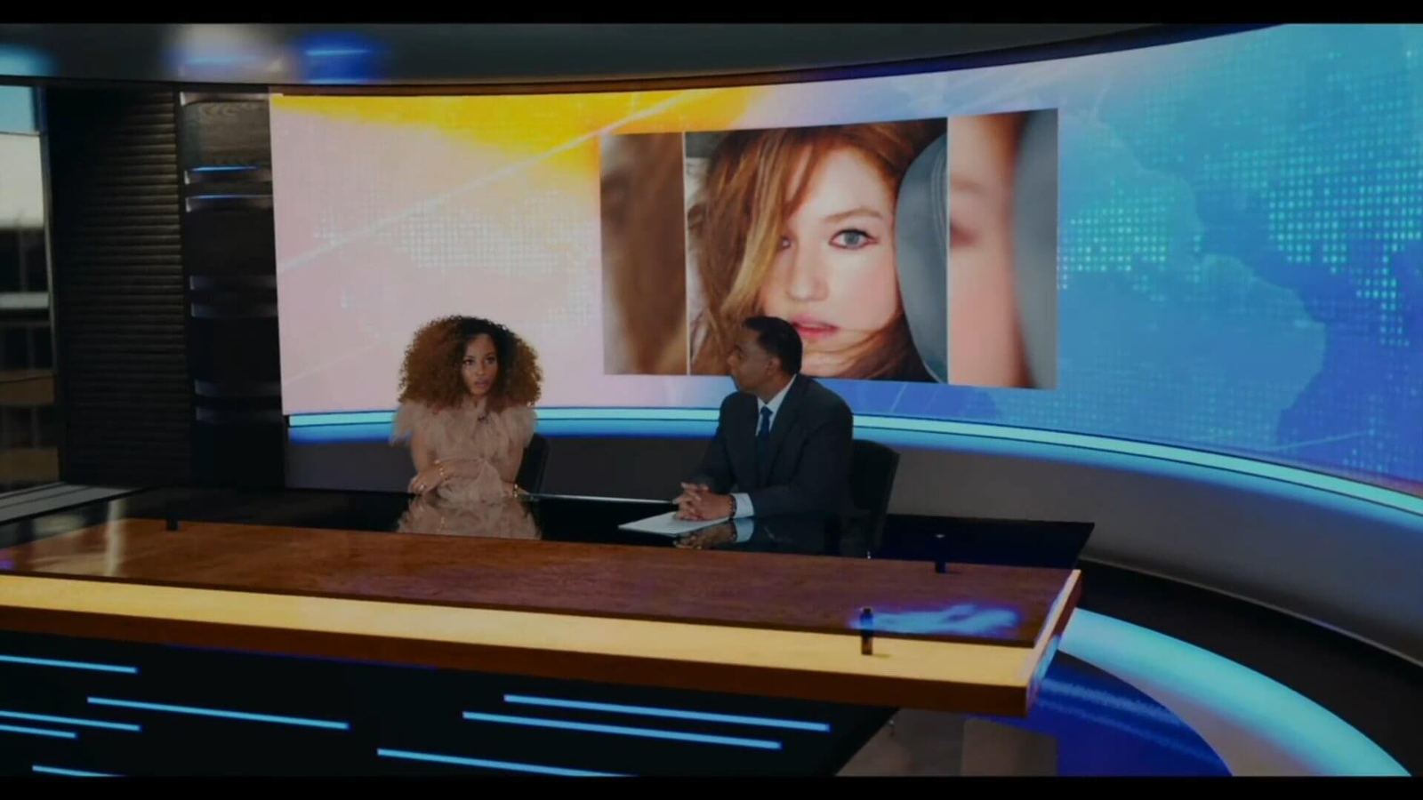 News set — two anchors at a wood desk before a curved video wall showing a fashion-portrait graphic, a virtual production set at L.A. & ABQ Castle Studios