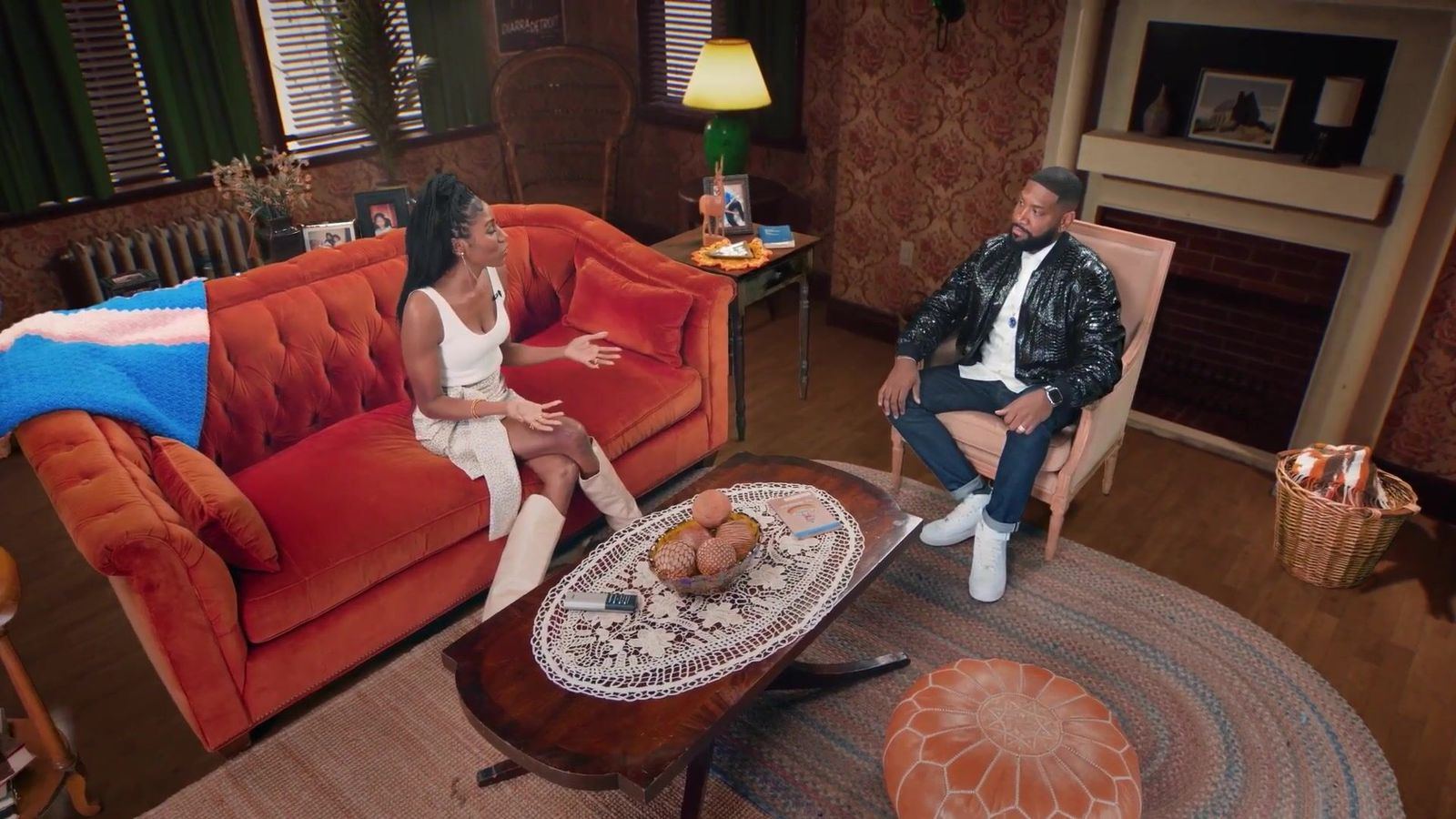 Press junket: a two-person interview on a living room virtual set with an orange sofa, a press junket sit-down format at L.A. & ABQ Castle Studios
