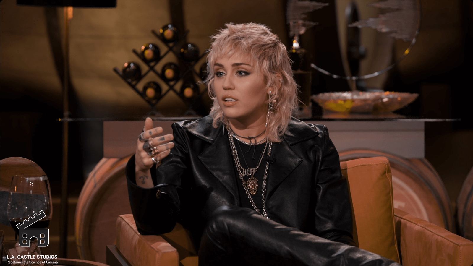 Talk show set — a guest in a black leather outfit gestures in an orange armchair within a warm lounge backdrop at L.A. & ABQ Castle Studios