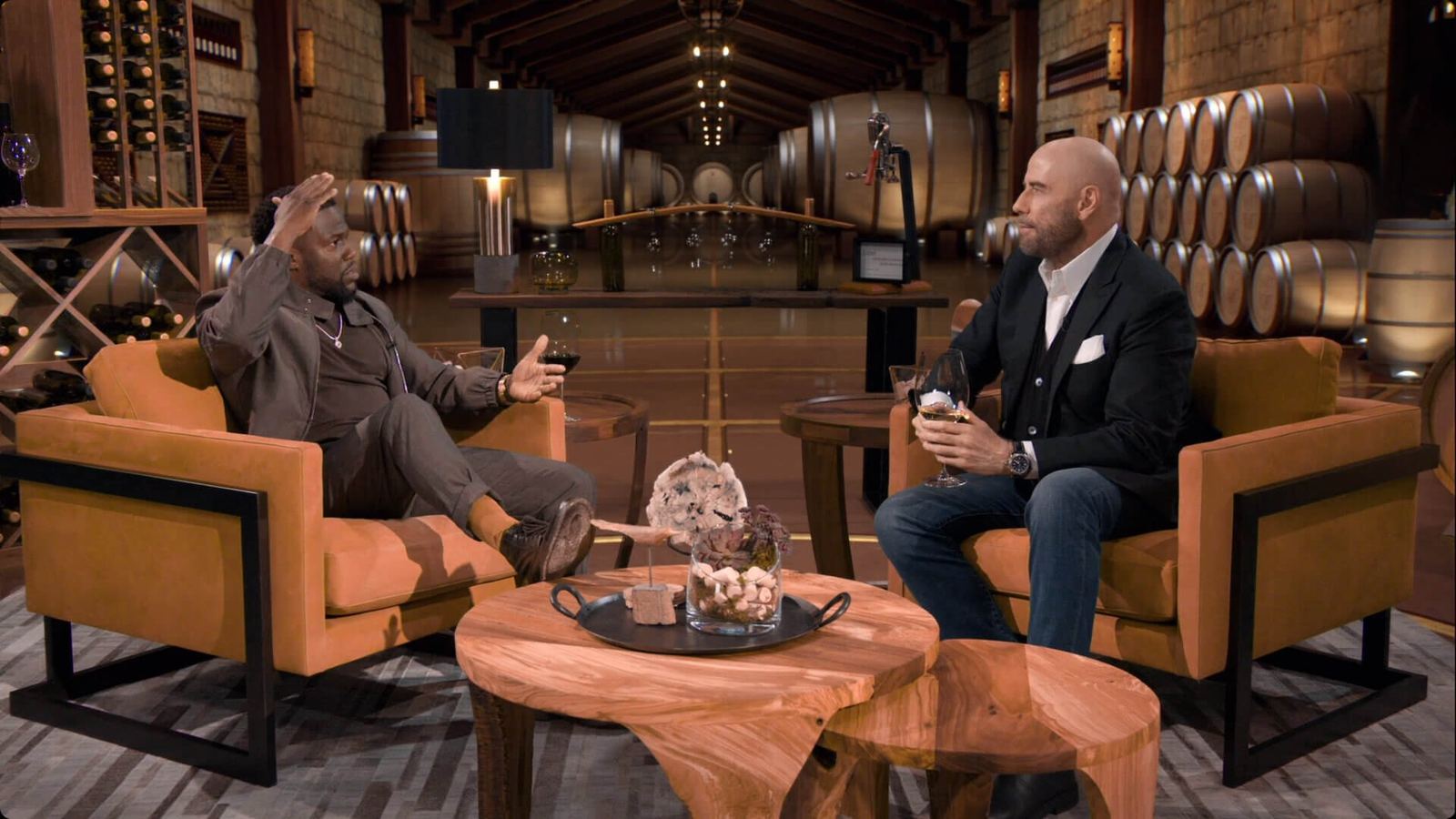 Host and guest in tan armchairs inside a virtual winery cellar lined with oak barrels, a talk show set at L.A. & ABQ Castle Studios