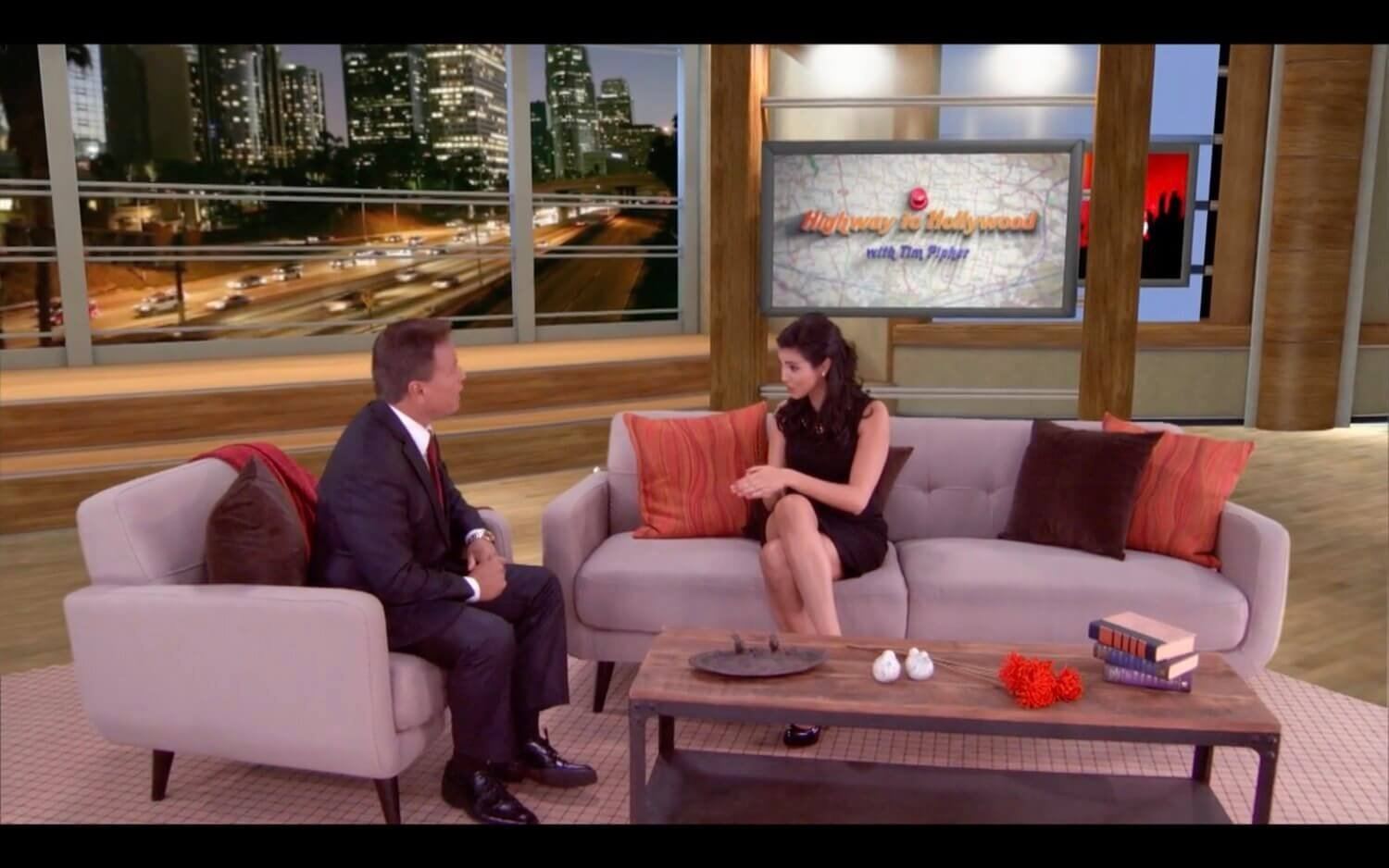 Talk show set — host and guest seated across a low table before a nighttime Los Angeles freeway and skyline backdrop, a virtual production set at L.A. & ABQ Castle Studios