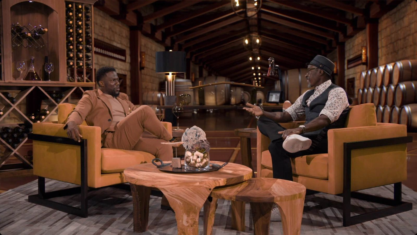 Talk show set — two guests converse in tan armchairs inside a virtual winery cellar with wine racks and oak barrels at L.A. & ABQ Castle Studios