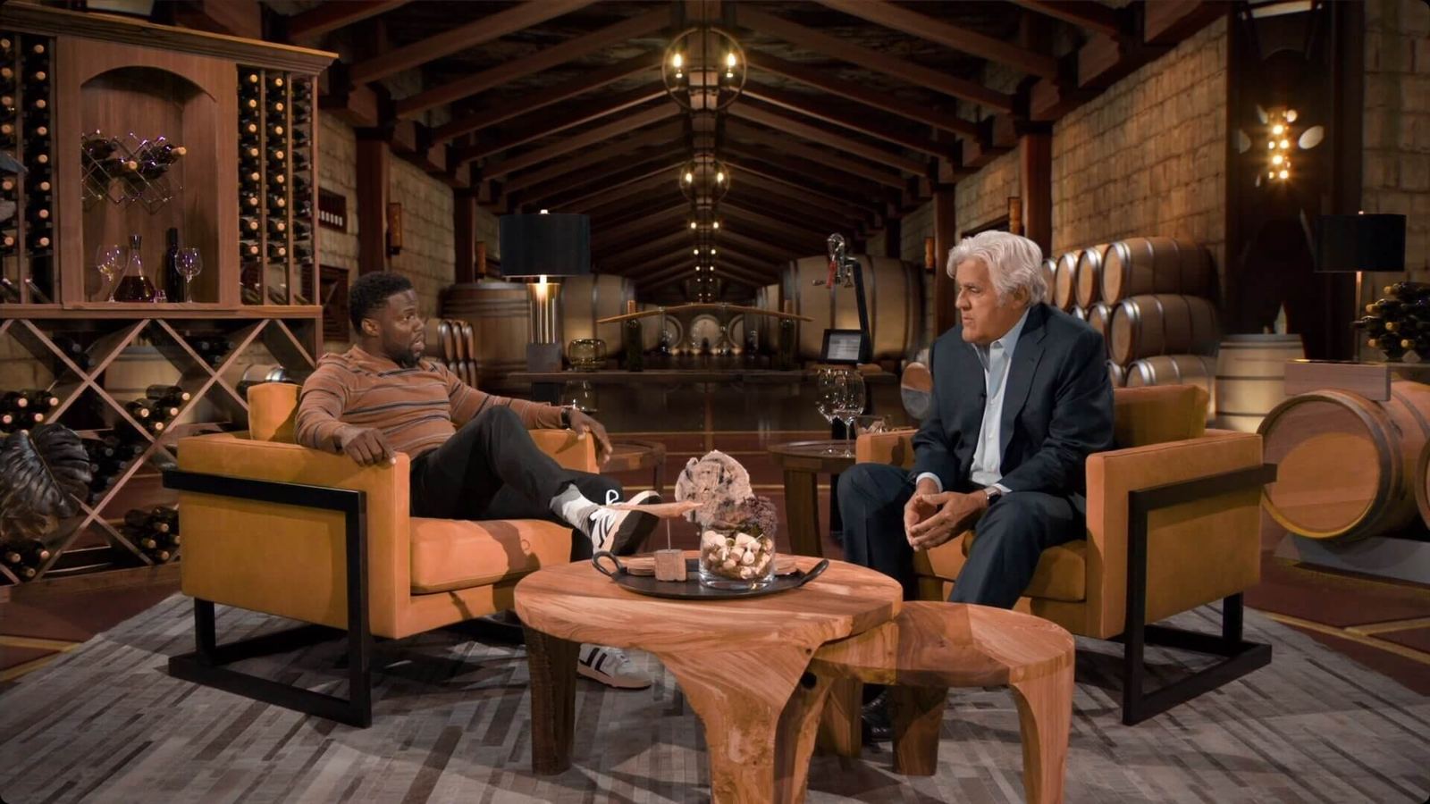 Talk show set: host and guest in tan armchairs inside a stone-walled virtual winery cellar with oak barrels at L.A. & ABQ Castle Studios